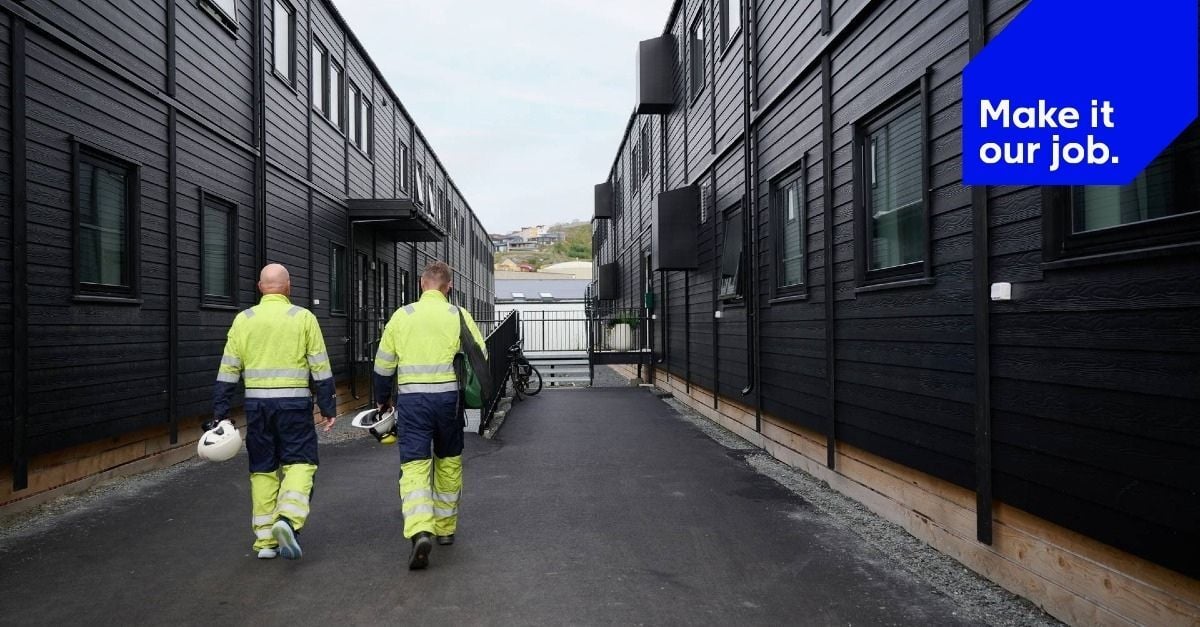 Adapteo-brändätty kuva, jossa kaksi huomiovaatteisiin pukeutunutta työntekijää kävelee mustien modulaaristen majoitusrakennusten välissä. Kuvassa on tunnuslause: “Make it our job.”
